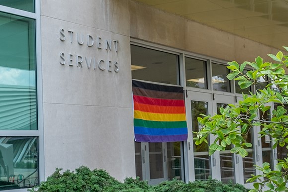 Flag made of horizontal strips of fabric in a rainbow pattern hanging next to a door outside the Student Services Building. 